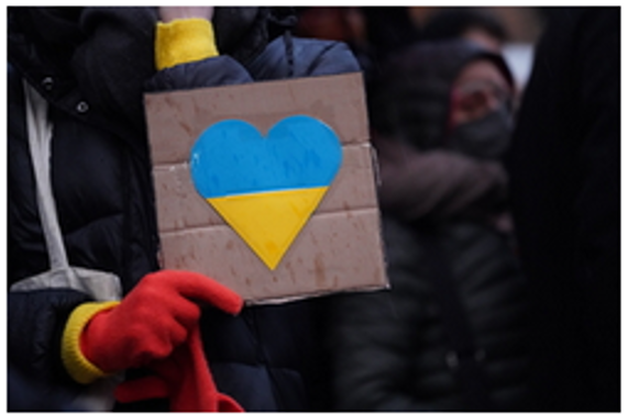 Auf einem Pappschild klebt ein Herz in den Farben der Ukraine - blau/rot, dieses Schild wird von jemanden in der Hand gehalten. Person hat roten Handschuh an.